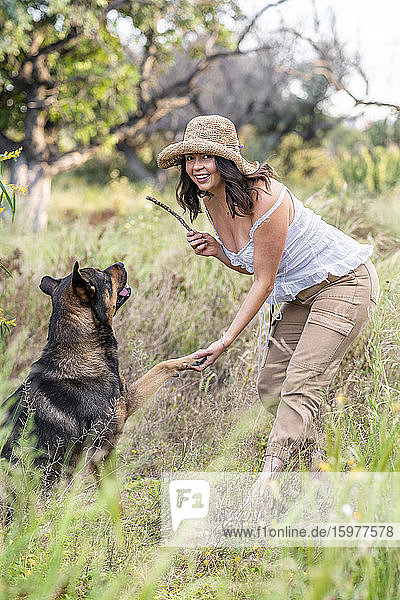 Porträt einer lächelnden schönen jungen Frau  die einen Stock hält  während sie mit einem Hund bei Pflanzen spielt  Alicante  Provinz Alicante  Spanien