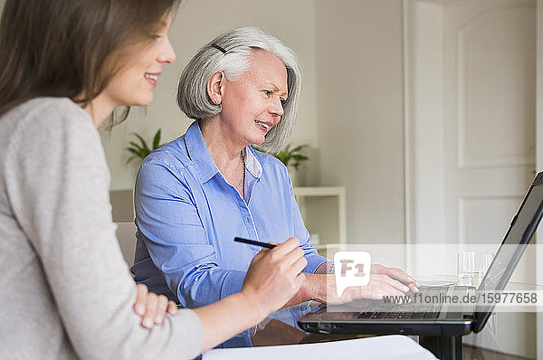 Porträt einer älteren Frau  die an einem Laptop arbeitet  während ihre erwachsene Tochter sie beobachtet