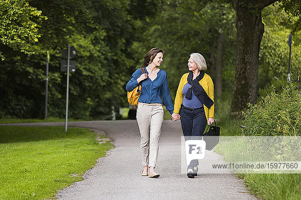 Lächelnde ältere Frau und erwachsene Tochter gehen Hand in Hand in einem Park und schauen sich an