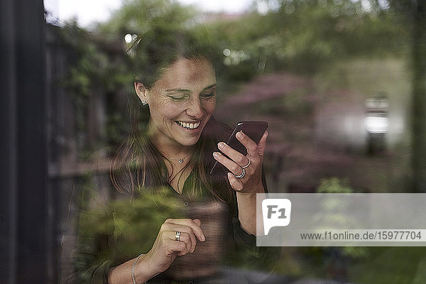 Lächelnde Frau im mittleren Erwachsenenalter  die zu Hause ein Mobiltelefon benutzt  gesehen durch ein Fenster