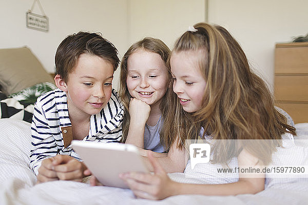 Porträt eines Jungen und seiner beiden Schwestern  die auf einem Bett liegen und ein digitales Tablet für den Hausunterricht benutzen