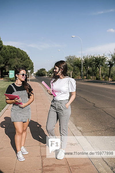 In voller Länge von lächelnden jungen Studentinnen  die Akten halten  während sie auf dem Fußweg auf dem Universitätscampus gegen den Himmel laufen