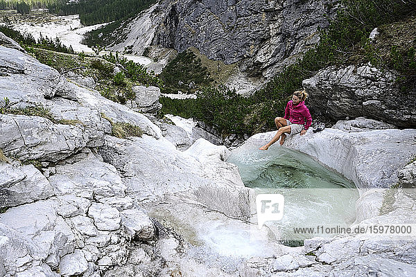 Ältere weibliche Touristin sitzt auf einem Felsen und spritzt Wasser im Bach