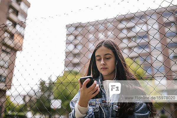 Niedriger Winkel Blick auf Teenager-Mädchen mit Smartphone  während gegen Maschendrahtzaun stehen
