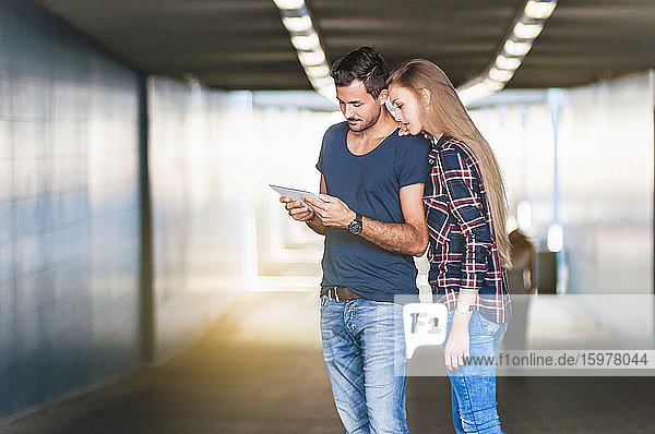 Junger Mann und junge Frau stehen in einer Unterführung und schauen auf ein digitales Tablet