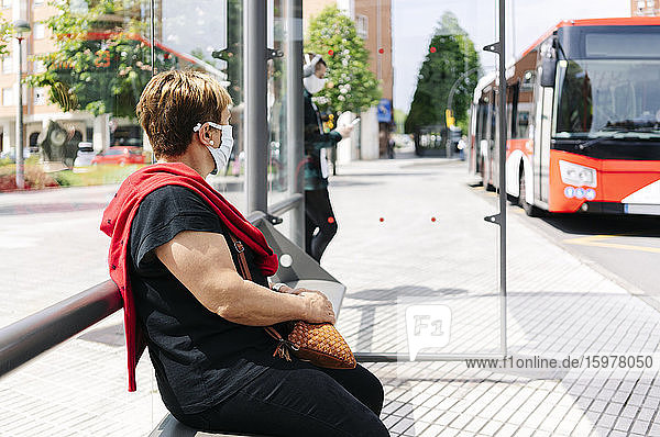 Ältere Frau mit Schutzmaske wartet an einer Bushaltestelle  Spanien