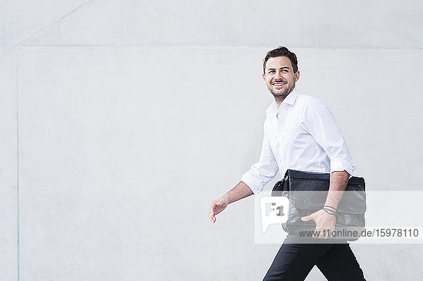 Smiling businessman with laptop bag walking along white wall