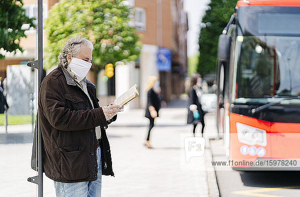 Älterer Mann mit Schutzmaske und Handschuhen liest ein Buch  während er an einer Bushaltestelle wartet  Spanien