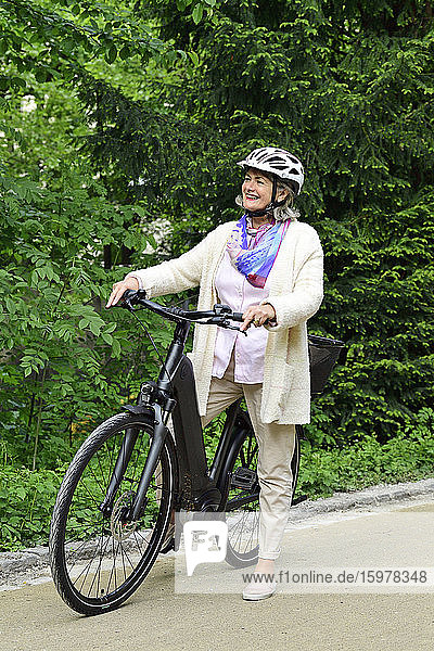 Glückliche ältere Frau mit Elektrofahrrad auf dem Fußweg im Park stehend