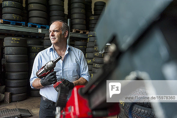 Ein älterer Unternehmer hält einen Schlagschrauber in der Hand  während er in einem Reifengeschäft wegschaut
