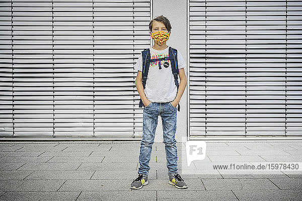 Boy with schoolbag wearing mask standing in front of building