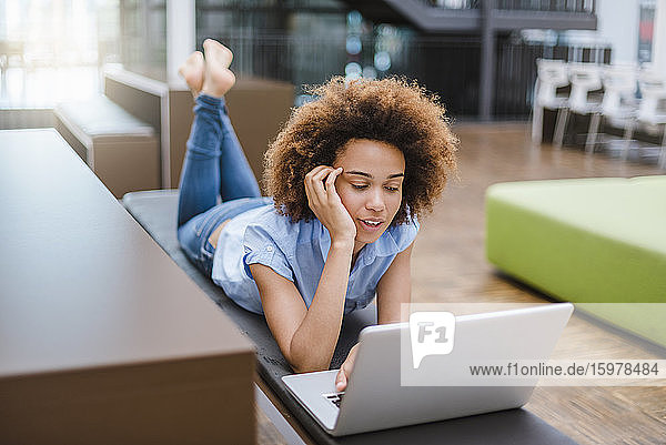 Junge Frau liegt auf einer Bank in einem modernen Büro und benutzt einen Laptop