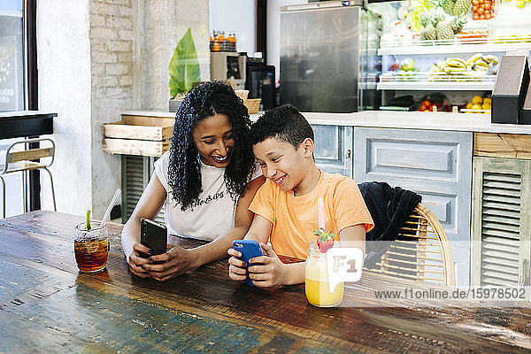 Fröhliche Mutter und Sohn benutzen Mobiltelefone am Tisch im Restaurant