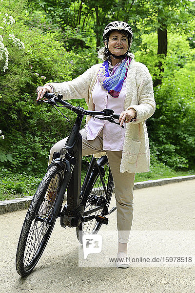 Nachdenkliche ältere Frau mit Elektrofahrrad auf dem Fußweg im Park stehend