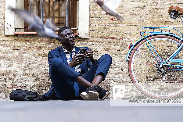 Junger Geschäftsmann mit Smartphone auf dem Boden sitzend in der Stadt