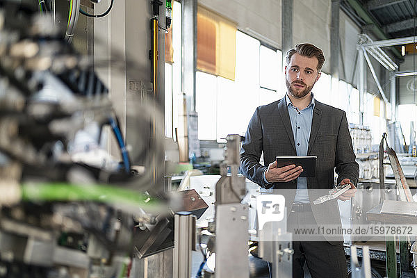 Young businessman with tablet and workpiece at a machine in a factory