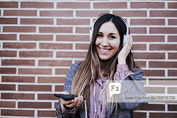 Portrait of happy woman listening music with headphones and smartphone in front of brick wall