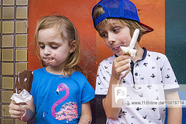 Niedliche Geschwister essen Eis am Stiel  während sie an der Wand stehen