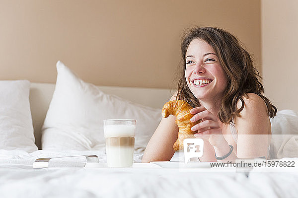 Happy young woman holding croissant while having breakfast in bed