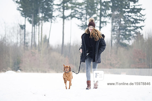 Smiling young woman walking with dog on snow against trees in forest