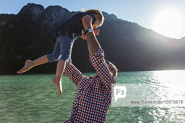 Vater hält seine Tochter hoch  während er vor einem Berg und einem See am Achensee steht  Bundesland Tirol  Österreich