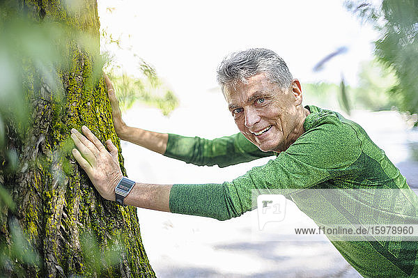 Portrait of smiling retired elderly man exercising while leaning on tree trunk at park
