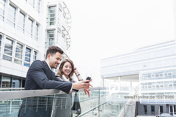 Ein Geschäftspaar benutzt ein Smartphone auf einem erhöhten Gehweg vor einem Bürogebäude