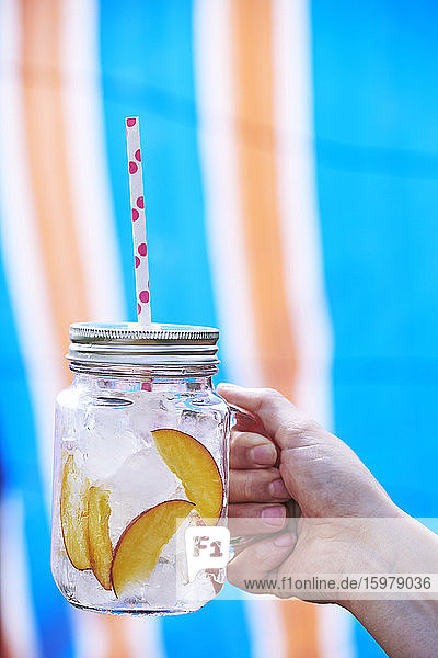Close-up of Woman's hand holding drink in mason jar filled with ice and slices of peach