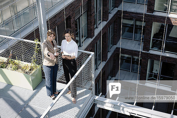 Zwei Geschäftsfrauen mit Tablet und Windradmodell bei einem Treffen auf dem Balkon eines Bürogebäudes