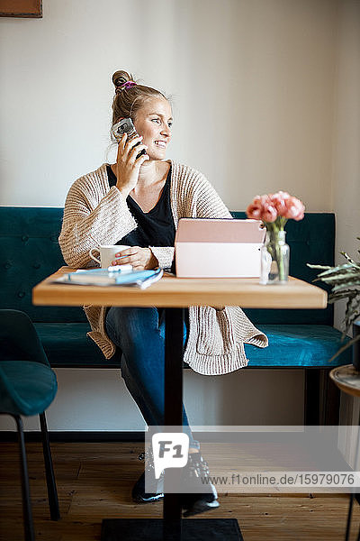 Lächelnde junge Frau am Telefon mit einer Tasse Kaffee und einem digitalen Tablet im Home Office