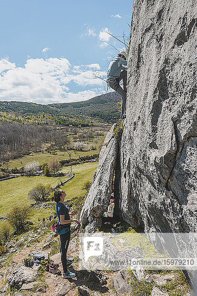 Young woman looking at man rock climbing against sky