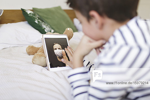 Boy lying on bed having video call on digital tablet with his mother wearng protective mask