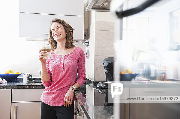 Glückliche Frau hält ein Glas Dalgona-Kaffee in der Hand  während sie zu Hause in der Küche steht