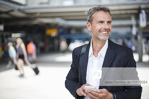 Lächelnder Geschäftsmann  der ein Smartphone hält und wegschaut  während er am Bahnhof steht