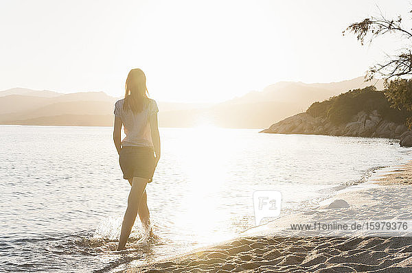 Rückenansicht einer Frau  die bei Sonnenuntergang am Meeresufer watet  Sardinien  Italien