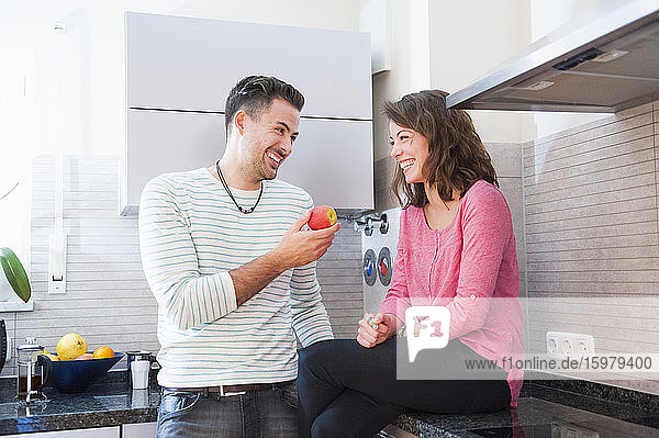 Happy man holding apple while looking at woman sitting on kitchen counter