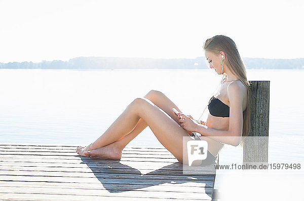 Junge Frau sitzt auf einem Steg vor einem See und hört mit Smartphone und Kopfhörern Musik