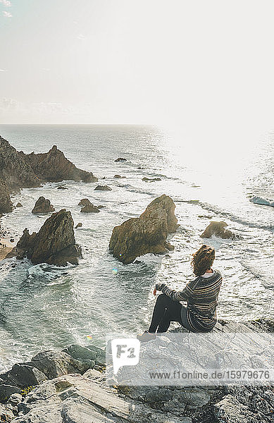 Junge Frau sitzt auf einer Felsformation und schaut auf das Meer  Praia da Ursa  Lisboa  Portugal