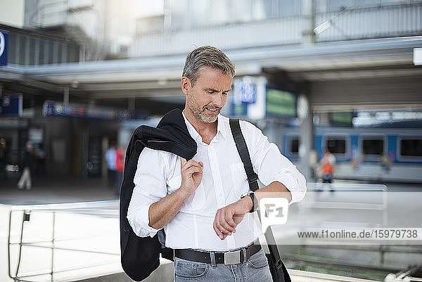 Businessman holding blazer checking time while standing against railroad station