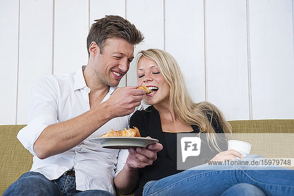 Smiling man feeding croissant to girlfriend while sitting on sofa at home