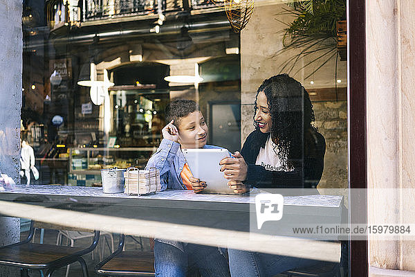 Lächelnder Junge sieht seine Mutter an  die mit einem digitalen Tablet in einem Restaurant sitzt  gesehen durch ein Fenster