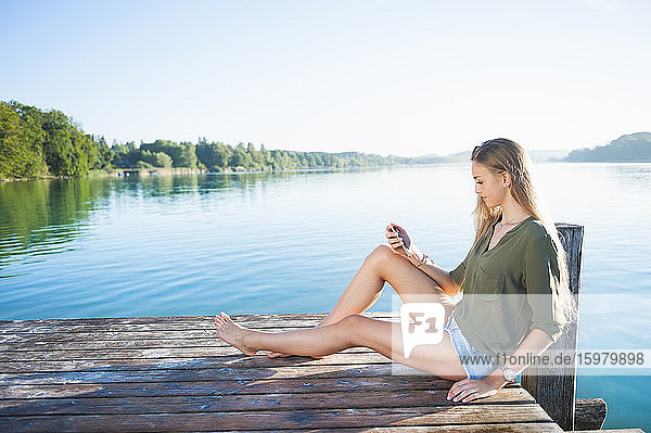 Junge Frau sitzt auf einem Steg am See und schaut auf ihr Handy