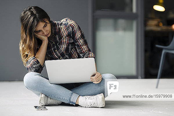Pensive young freelancer sitting on the floor looking at laptop