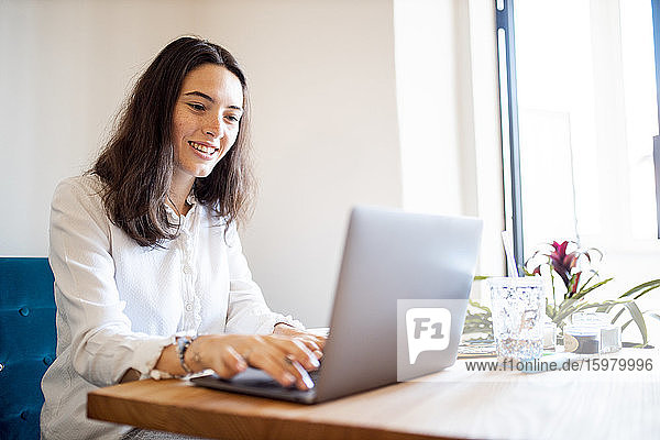 Porträt einer lächelnden jungen Frau  die im Büro zu Hause am Laptop arbeitet