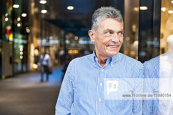 Porträt eines lächelnden älteren Mannes  der sich an eine Glasfront in einem Einkaufszentrum lehnt