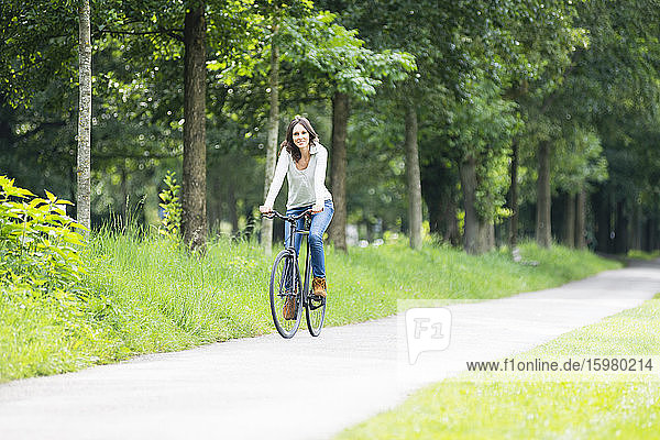 Lächelnde Frau auf dem Fahrrad auf dem Fußweg gegen grüne Bäume im Park