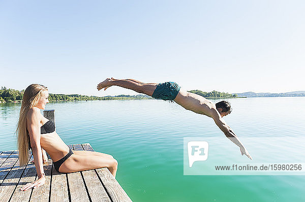 Young man in swimming shorts jumping from jetty into lake while his girlfriend watching him