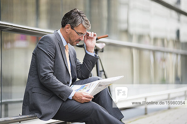 Businessman wearing eyeglasses reading newspaper while sitting on railing