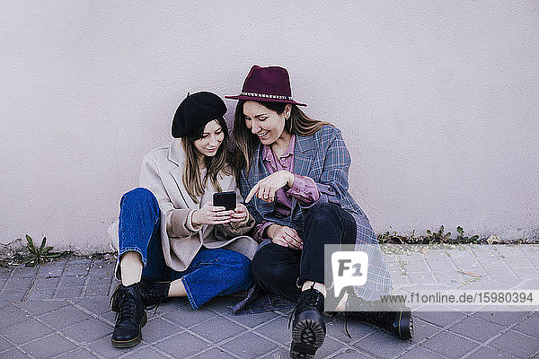 Porträt von zwei glücklichen Frauen  die zusammen auf dem Bürgersteig sitzen und auf ihr Handy schauen