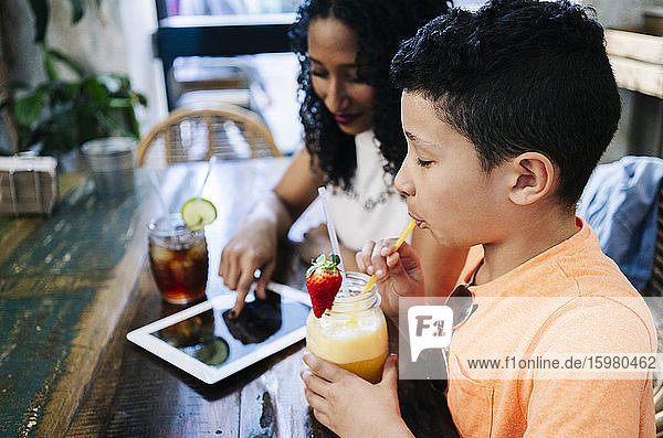 Side view of boy enjoying smoothie by mother sitting with digital tablet at restaurant